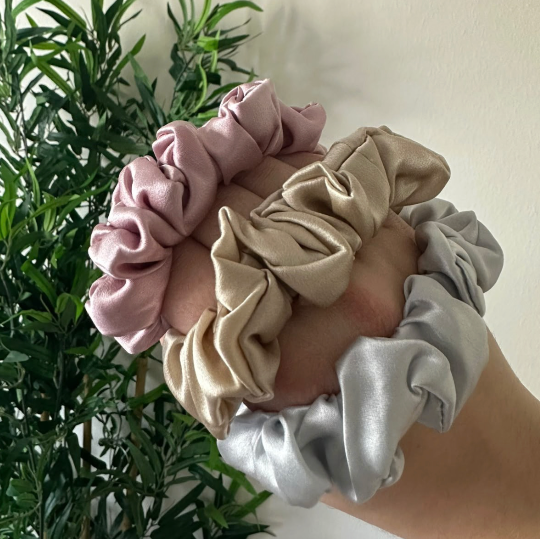 Trio de chouchous en soie rose poudré, beige et gris clair tenu en main devant une plante