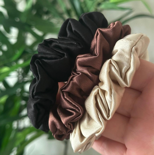 Trio de chouchous en soie noir, marron chocolat et creamy beige tenu en main devant une plante