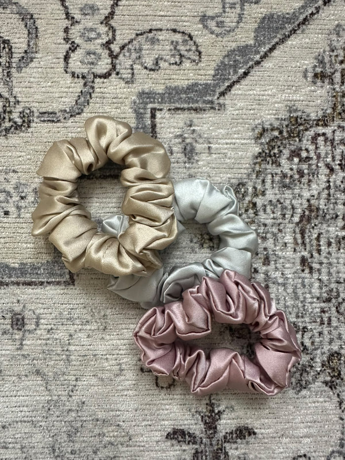 Trois chouchous en soie beige, gris clair et rose poudré posés sur un tapis